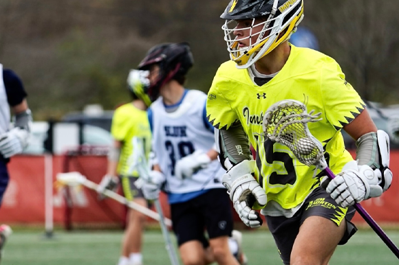 Person playing lacrosse in a yellow jersey with a blurred background of other players and equipment.