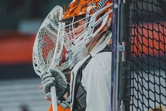 Person wearing a lacrosse helmet and holding a goalie lacrosse stick behind a goal.